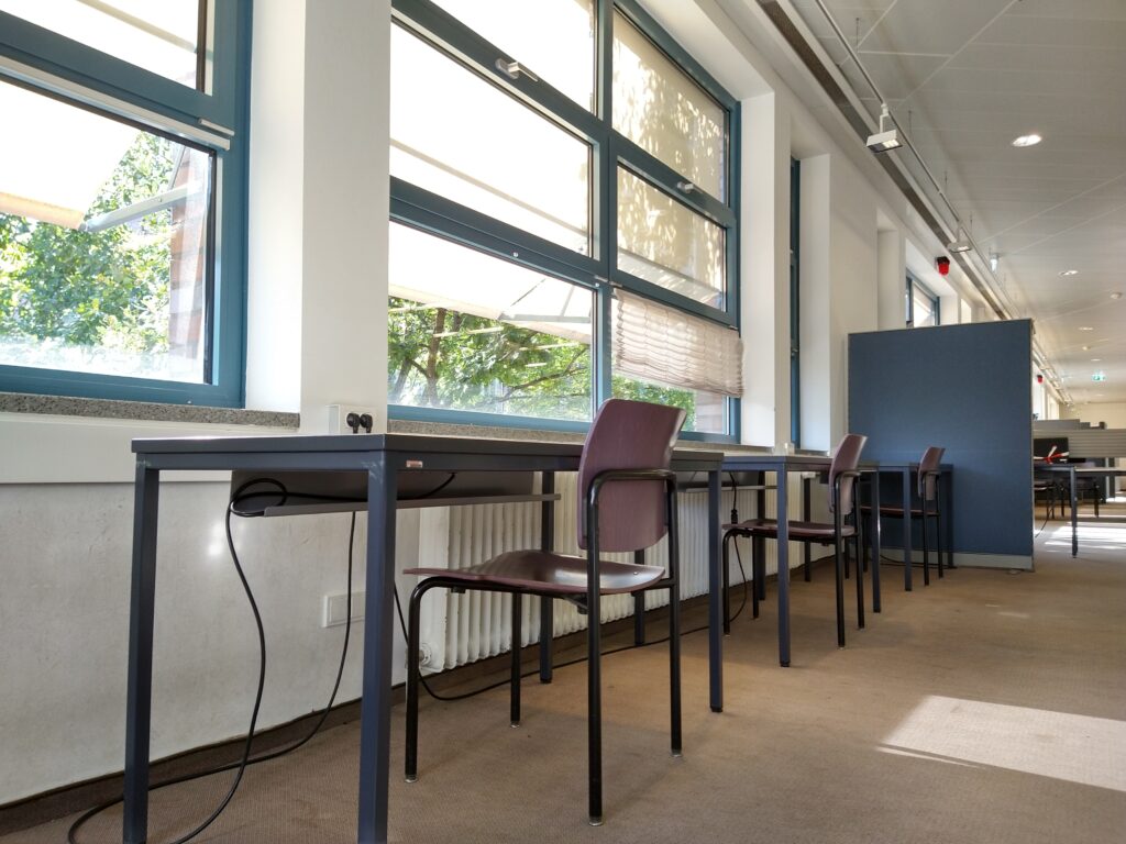 Eine Reihe Arbeitstische mit Stühlen vor einer Fensterwand.