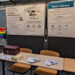 Foto des Stands der Campus-Austellung. Zu sehen sind drei Plakate an einer Stellwand, die auf Bibliotheksausweis, Escape Game und Open Library hänge. Davor steht ein Tisch, auf dem Flyer liegen und eine kleine Schatzkiste steht.