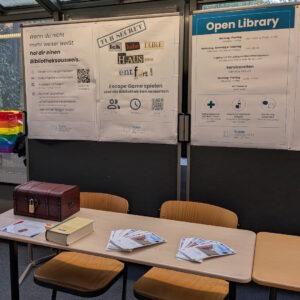 Foto des Stands der Campus-Austellung. Zu sehen sind drei Plakate an einer Stellwand, die auf Bibliotheksausweis, Escape Game und Open Library hänge. Davor steht ein Tisch, auf dem Flyer liegen und eine kleine Schatzkiste steht.